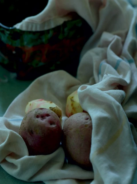 Close-up still life of whole and peeled red potatoes with a white cloth, and a glimpse of a black bowl in dim, moody lighting.