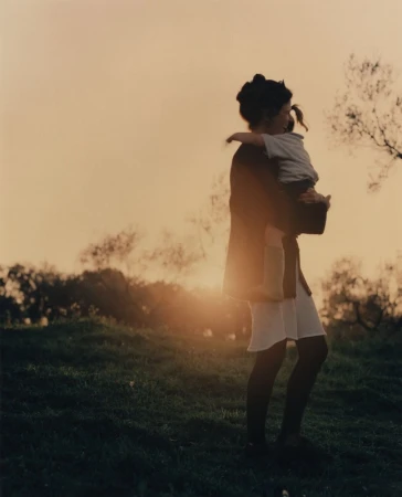 An adult holding a child in silhouette against a golden sunset sky over a grassy hill.