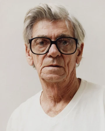 Portrait of an elderly man with grey hair wearing black eyeglasses and a white t shirt against a plain background