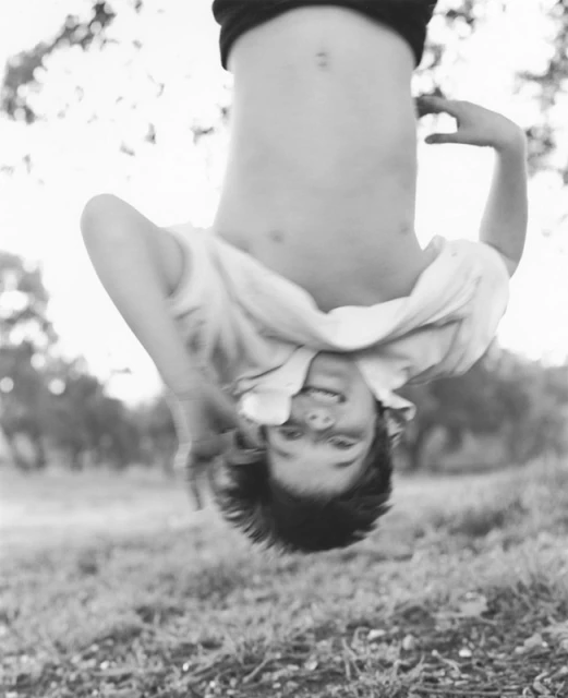 Black and white image of a smiling child hanging upside down outdoors with a blurred grassy field and trees in the background.