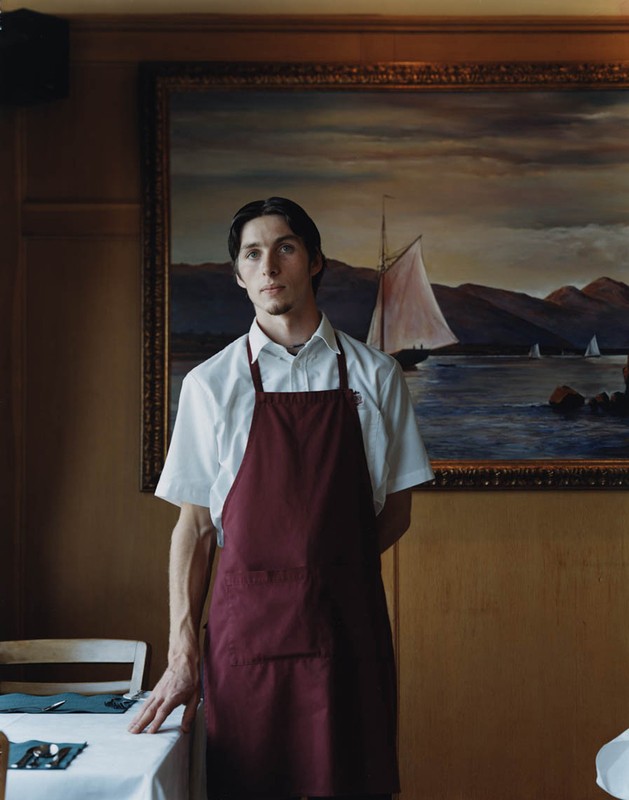 Portrait of a waiter standing in a restaurant, wearing a maroon apron and collared shirt, posed in front of a painting of sailboats.