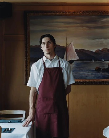 Portrait of a waiter standing in a restaurant, wearing a maroon apron and collared shirt, posed in front of a painting of sailboats.
