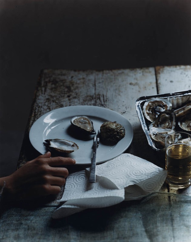Still life photo on a weathered table with oysters on a plate, an aluminum tray, a knife, a glass of liquid, and a hand.
