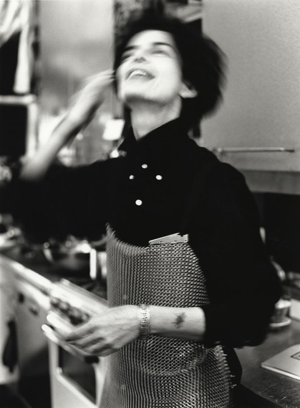 Black-and-white image of a smiling adult female wearing a chef's apron in a kitchen. Motion blur is visible, giving the photo a dynamic feel.