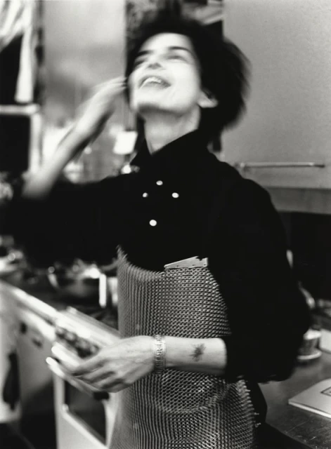 Black-and-white image of a smiling adult female wearing a chef's apron in a kitchen. Motion blur is visible, giving the photo a dynamic feel.