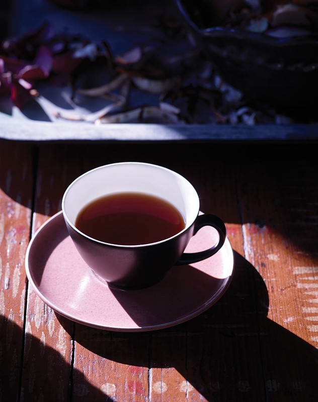 Elevated view of black tea cup with pink saucer on weathered wood surface, bathed in sunlight and shadow, dark floral background.