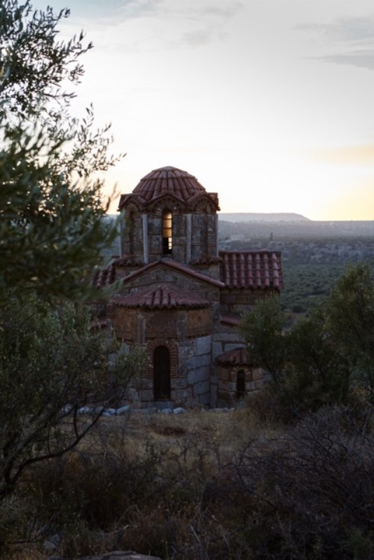 Exterior daytime view of a stone church with a red tile roof and dome surrounded by trees in a rural landscape with a sky.