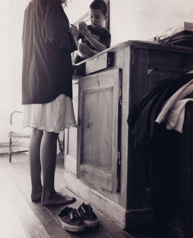 Black and white photograph features a child on a counter while another person stands nearby, with shoes on the wooden floor.