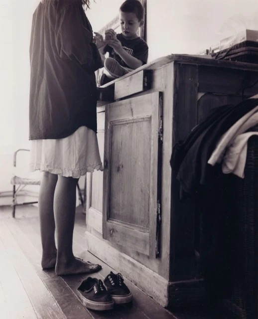 Black and white photograph features a child on a counter while another person stands nearby, with shoes on the wooden floor.