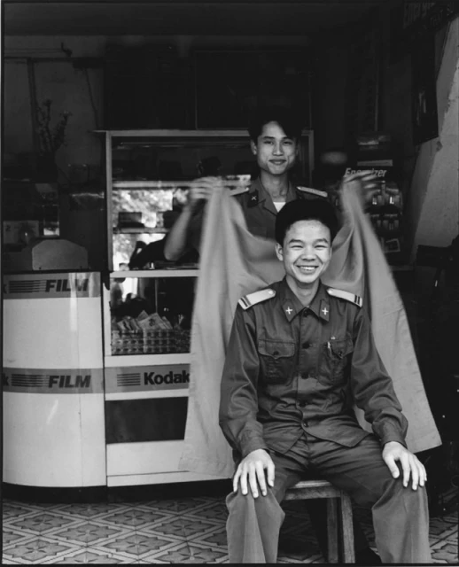 Two smiling men in uniform pose indoors with a film and camera display. One sits while the other holds a garment behind him.