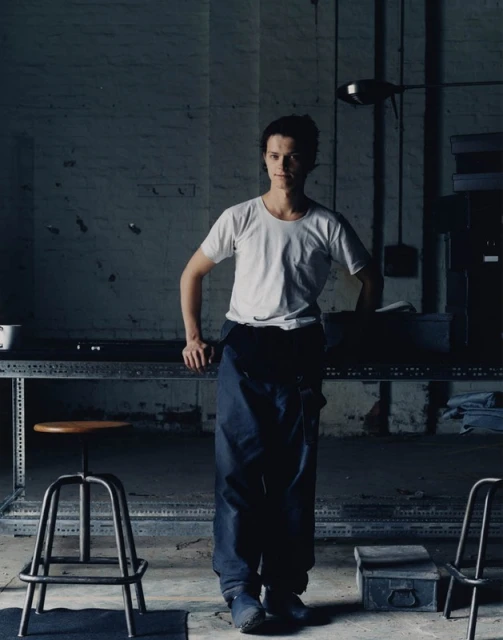 Full body shot of a young adult standing in an industrial setting near a metal table and stools wearing an apron and t-shirt.