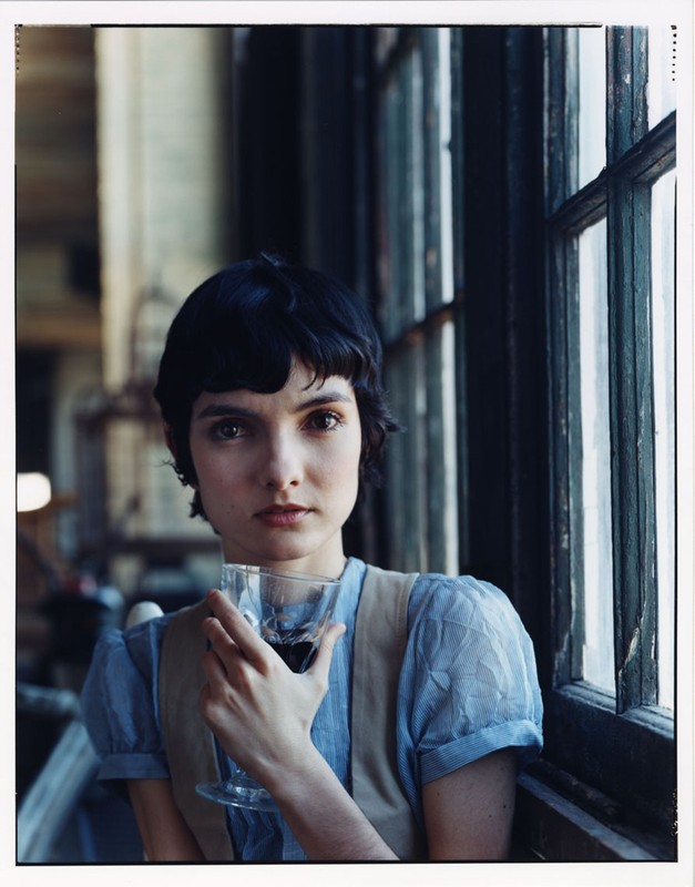 Portrait of a woman with short dark hair in front of a window, holding a stemmed glass of red wine, wearing a blue blouse and vest.