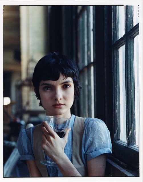 Portrait of a woman with short dark hair in front of a window, holding a stemmed glass of red wine, wearing a blue blouse and vest.