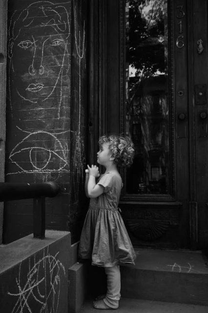 Black and white shot of child in dress standing in front of door, looking at white chalk drawings of face and eye on the wall.
