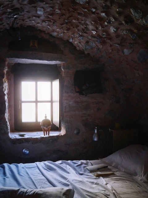 Bedroom interior featuring a bed with striped linens, a window letting in bright light, stone walls, and an antique ceramic object.