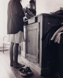 Black and white portrait of a woman standing barefoot near a wooden cabinet where a young boy is seated.