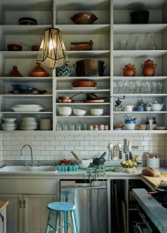 A rustic kitchen with white open shelving filled with pottery, glassware, and cookware above a stainless steel dishwasher and blue stool.