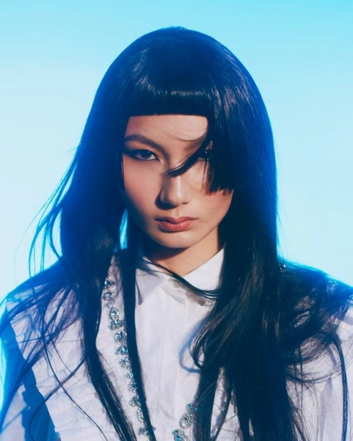 Headshot of a fashionable young woman with a modern black bob haircut, wearing a white shirt, against a gradient light blue background.