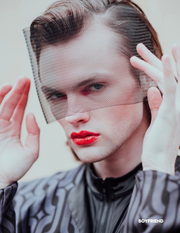 Headshot of a male with red lips and a black net veil. He wears a patterned shirt with a high neck and has light skin.