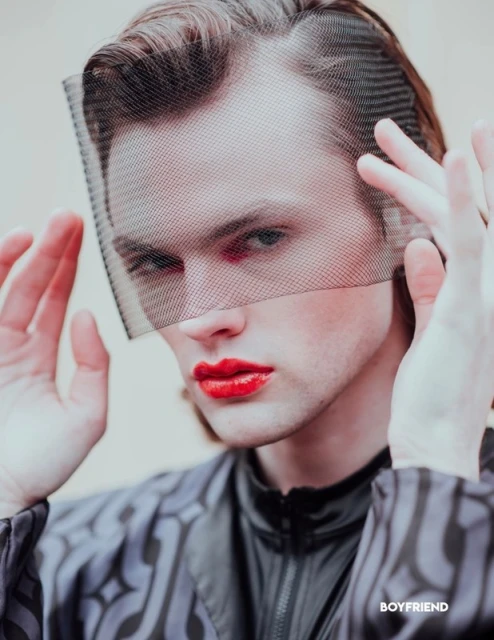 Headshot of a male with red lips and a black net veil. He wears a patterned shirt with a high neck and has light skin.