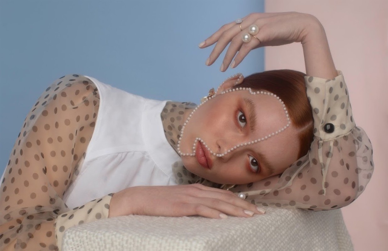 Portrait of an adult female with red hair, wearing a white top and sheer polka dot blouse, posing with pearls on her face.