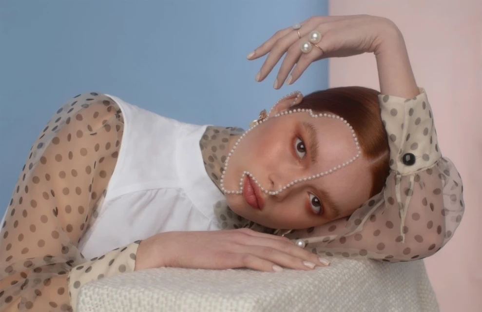 Portrait of an adult female with red hair, wearing a white top and sheer polka dot blouse, posing with pearls on her face.