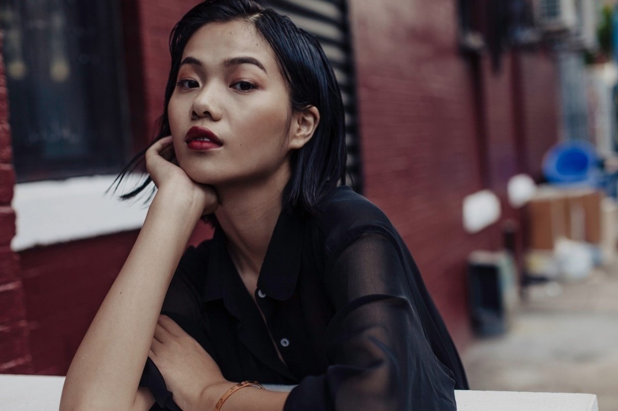 Close-up portrait of a young Asian woman with dark hair and red lipstick, wearing a sheer black blouse, posing outdoors against a red brick wall.