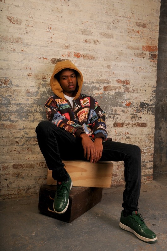 A young adult male in a hooded jacket sits on wooden crates against a brick wall, showcasing streetwear fashion and urban style.