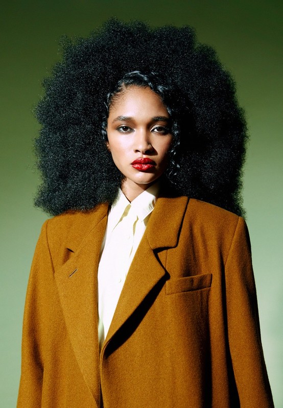 Portrait of a Black woman with a large afro, wearing a mustard-colored coat and white collared shirt, set against a green background.