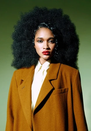Portrait of a Black woman with a large afro, wearing a mustard-colored coat and white collared shirt, set against a green background.