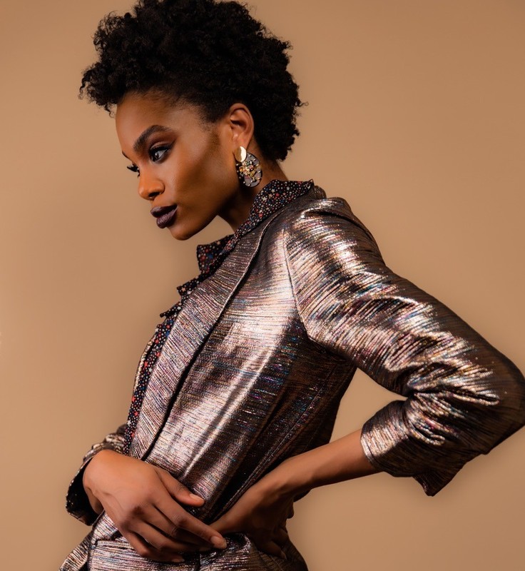 An adult woman poses indoors wearing a metallic gold blazer, with her hands on her hips, dark lipstick, and dangling earrings.