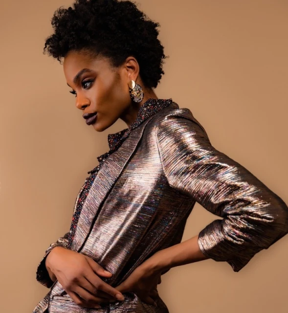 An adult woman poses indoors wearing a metallic gold blazer, with her hands on her hips, dark lipstick, and dangling earrings.