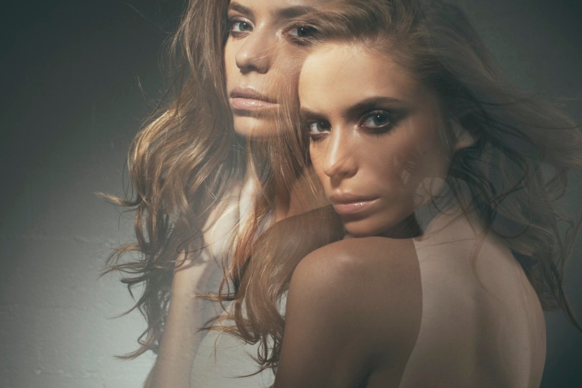 Portrait of two adult females with long wavy hair, one looking at the camera in a photo studio with soft lighting and a dark background.