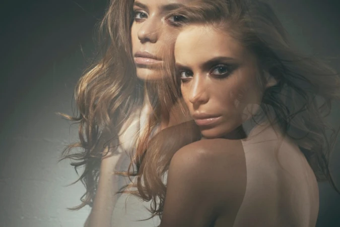 Portrait of two adult females with long wavy hair, one looking at the camera in a photo studio with soft lighting and a dark background.