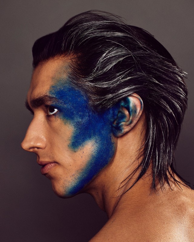 Profile view of a male model with dark hair and blue face paint, creating a bold and artistic beauty statement against a grey backdrop.