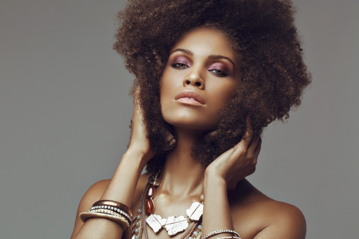 Portrait of a woman with a large afro hairstyle, wearing statement necklaces and bracelets against a plain grey background
