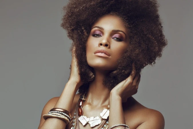 Portrait of a woman with a large afro hairstyle, wearing statement necklaces and bracelets against a plain grey background