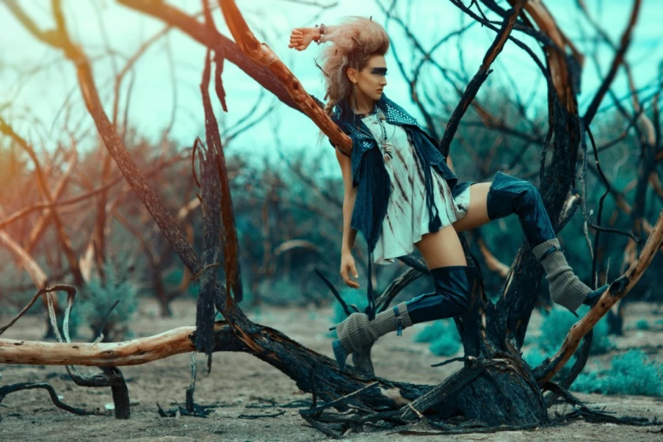 Woman in avant-garde fashion posing in a desolate landscape, wearing thigh-high boots and edgy makeup, amidst sparse vegetation.