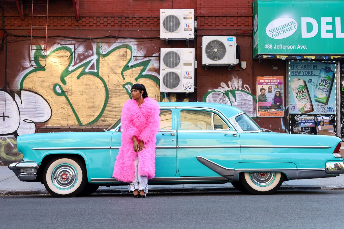 An adult female wearing a pink coat poses next to a light blue vintage car with white-wall tires on a city street with graffiti.