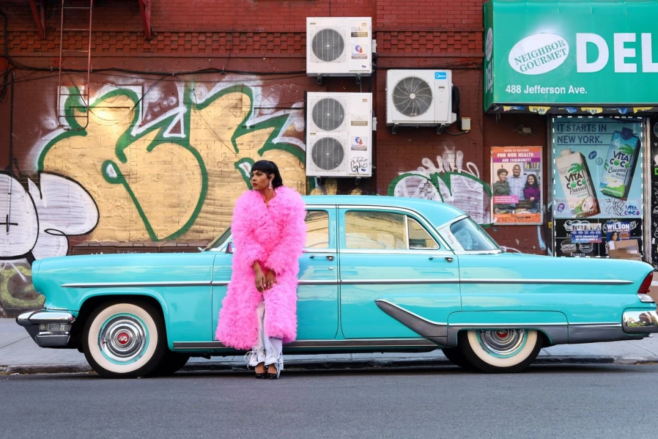 An adult female wearing a pink coat poses next to a light blue vintage car with white-wall tires on a city street with graffiti.