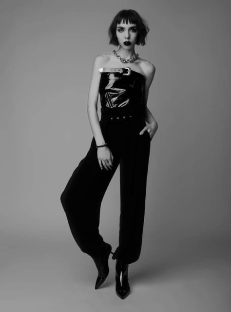 A full shot in black and white shows a slender adult woman with dark hair and dark lipstick wearing a shiny black top and black pants.