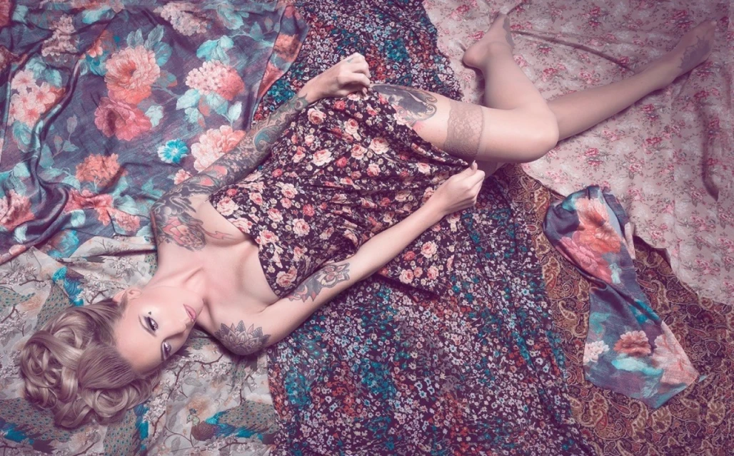 Woman lying on patterned fabrics wearing a floral dress, sheer hosiery and tattoos, in a studio environment with soft lighting.