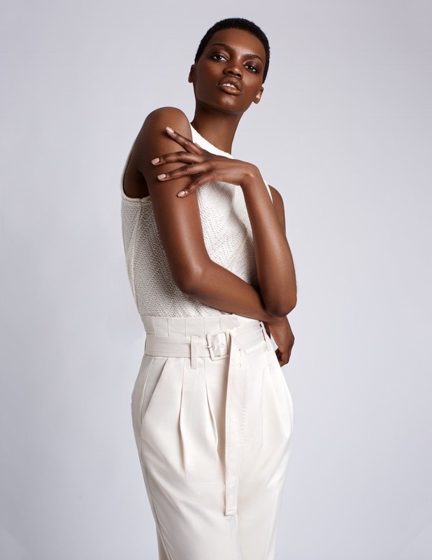 Black woman with short hair wearing a textured white sleeveless top and high-waisted cream trousers against a white background