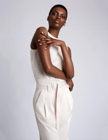 Black woman with short hair wearing a textured white sleeveless top and high-waisted cream trousers against a white background