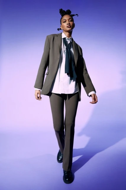 A full shot of an adult female fashion model walking towards the camera, wearing a suit and tie against a graduated purple background.