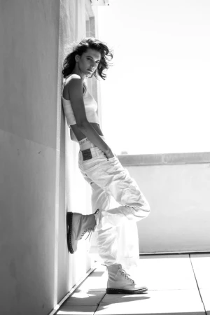 Model leaning against a wall outdoors wearing a white crop top, white cargo pants, and boots, styled with make-up by Marvin Alexander.