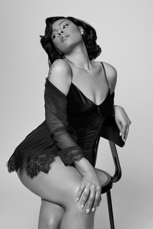 Black and white portrait of a woman in a black negligee sitting on a chair against a white backdrop, looking up and to the side.