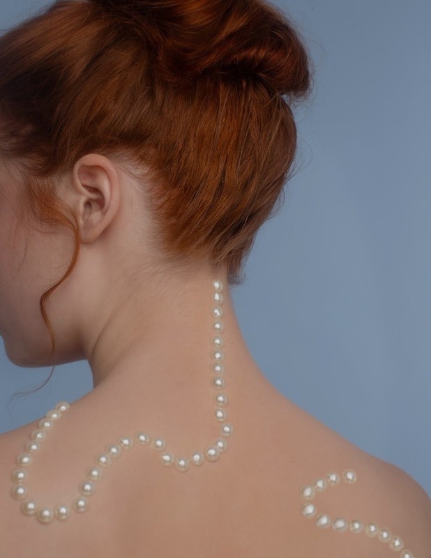 Back view of a redhead woman with pearls draped on her skin against a light blue background in a beauty and fashion portrait.