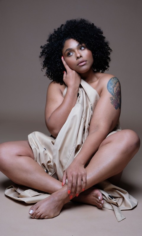 Portrait of a Black woman with curly hair, draped in beige fabric, sitting with crossed legs in a studio with a neutral background.