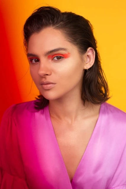 Close-up portrait of a young woman with bold red eyeshadow, fair skin, and a pink satin dress against a vibrant orange and red background.